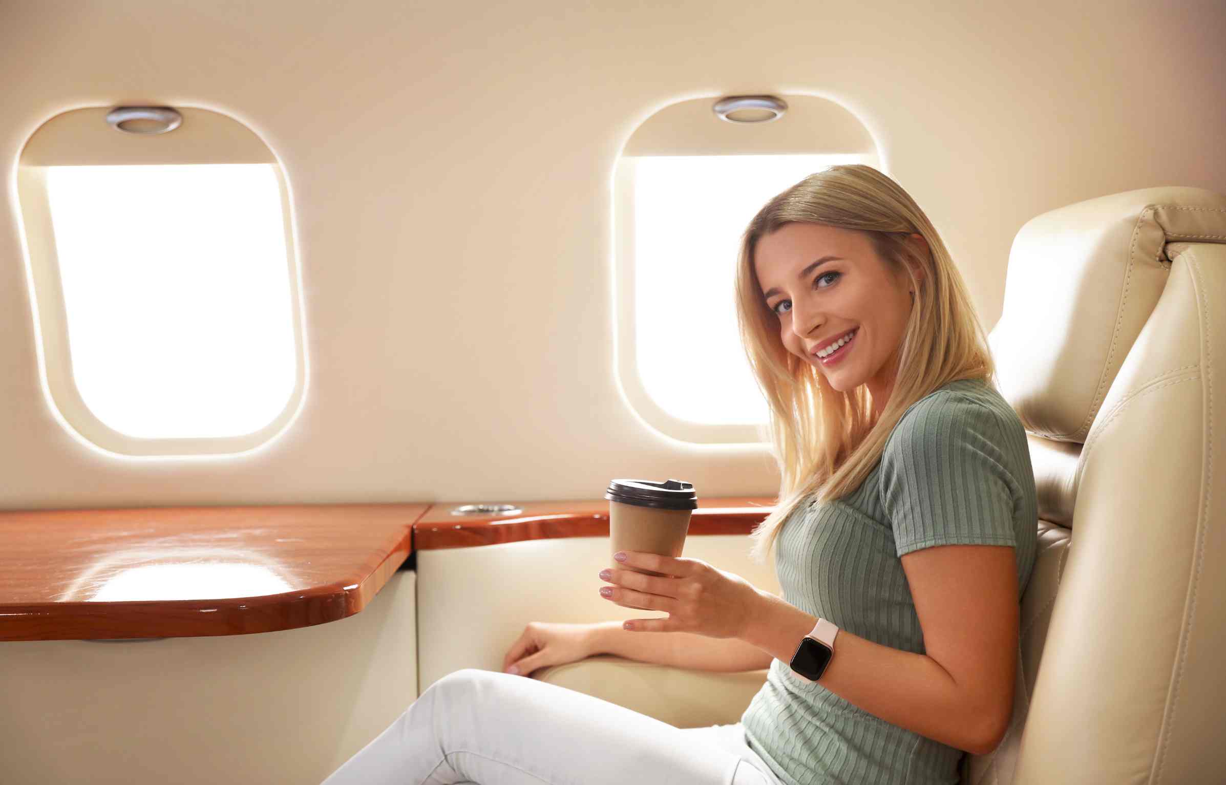 Smiling woman seated in a luxury cabin holding a paper coffee cup, enjoying a relaxed flight in a leather seat.