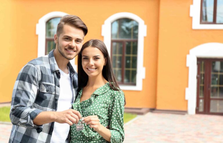 A happy young couple stands in front of their new house, smiling and holding a key.