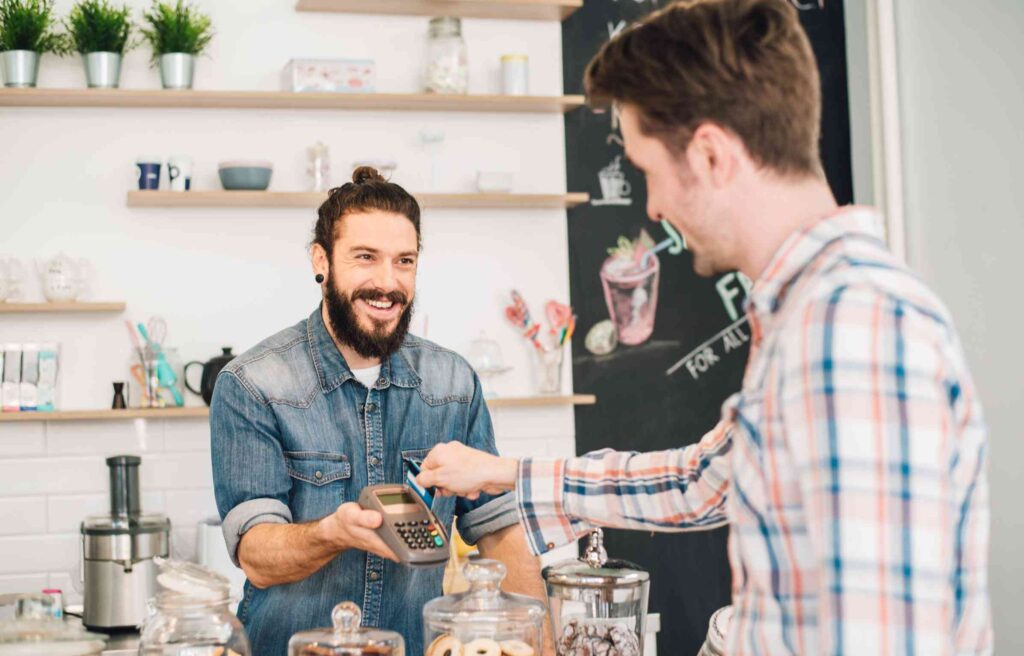 A smiling shopkeeper holds a card reader as a customer slides the new united business card to make a payment at the counter.