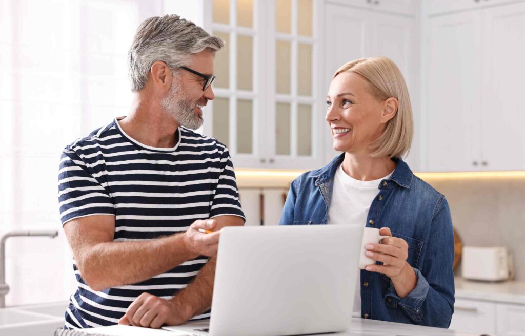 Smiling middle-aged couple planning finances on a laptop, discussing retirement withdrawal bucket strategy at home.