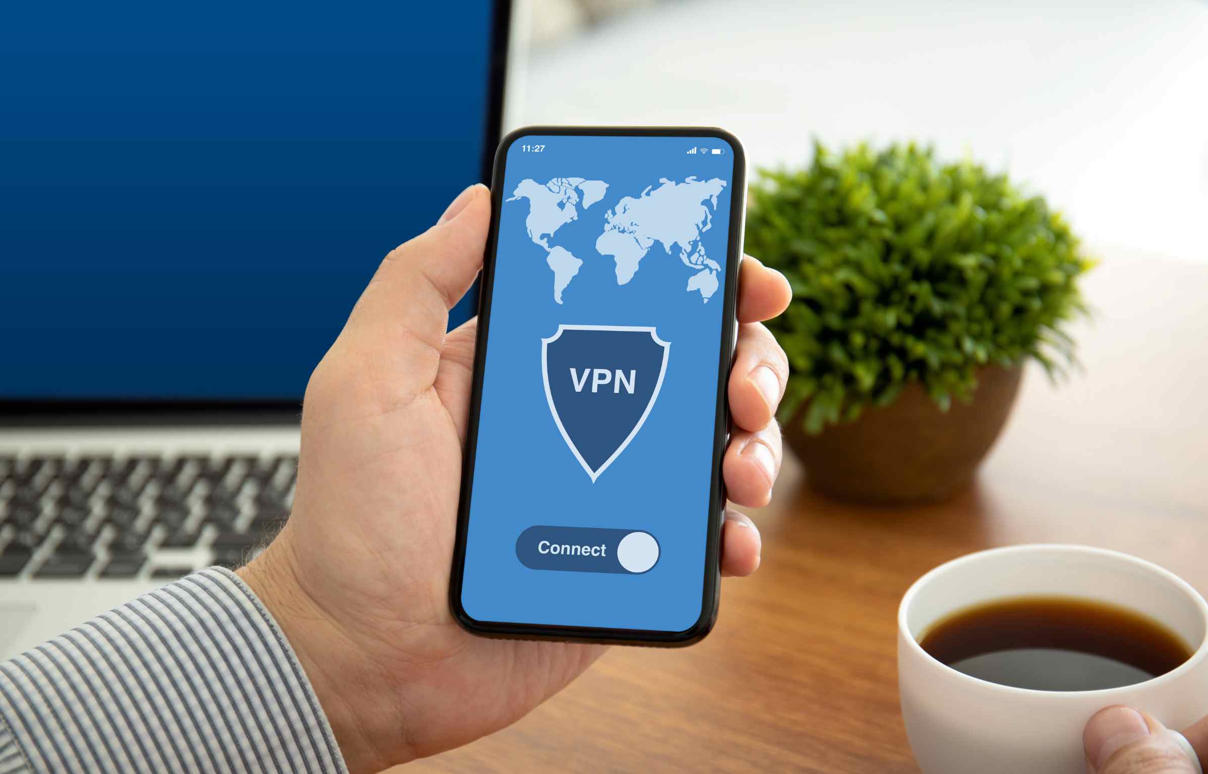 A person holding a smartphone with a VPN app open, showing a world map and connect button, while sitting at a desk with a laptop and a cup of coffee.