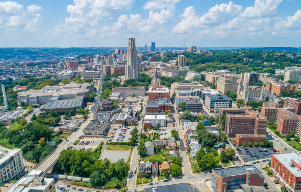 Aerial view of Pittsburgh, Pennsylvania with downtown buildings, green hills, and the Cathedral of Learning visible in the skyline.