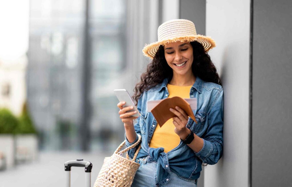 Smiling traveler holding a passport and boarding pass while using the amex travel app on her smartphone.