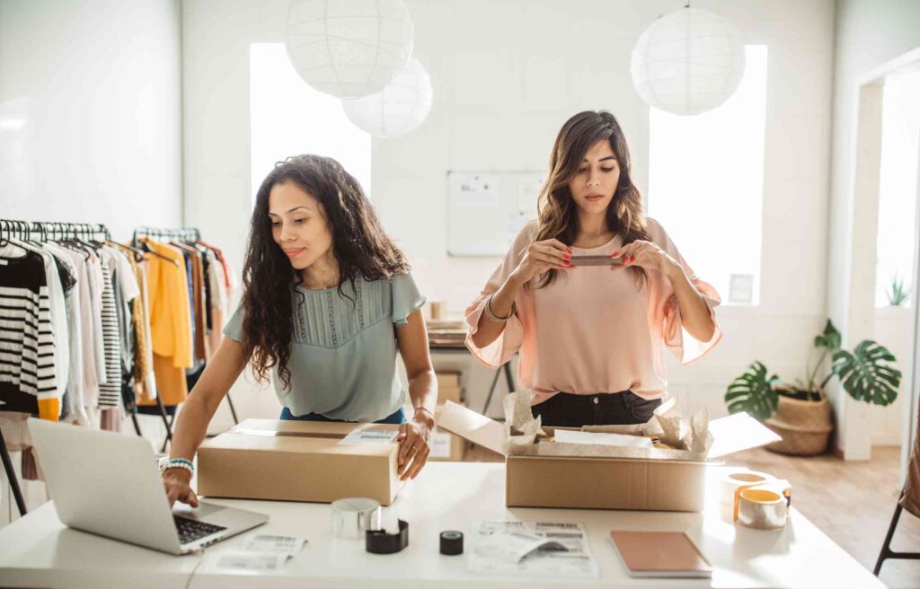 Two small business owners working together in a clothing shop, one packing products into boxes while the other uses a laptop.