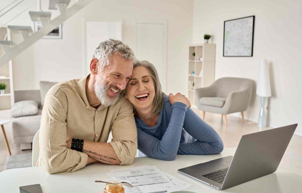 A cheerful middle-aged couple laughs together while planning retirement finances on a laptop, showing comfort and emotional well-being.