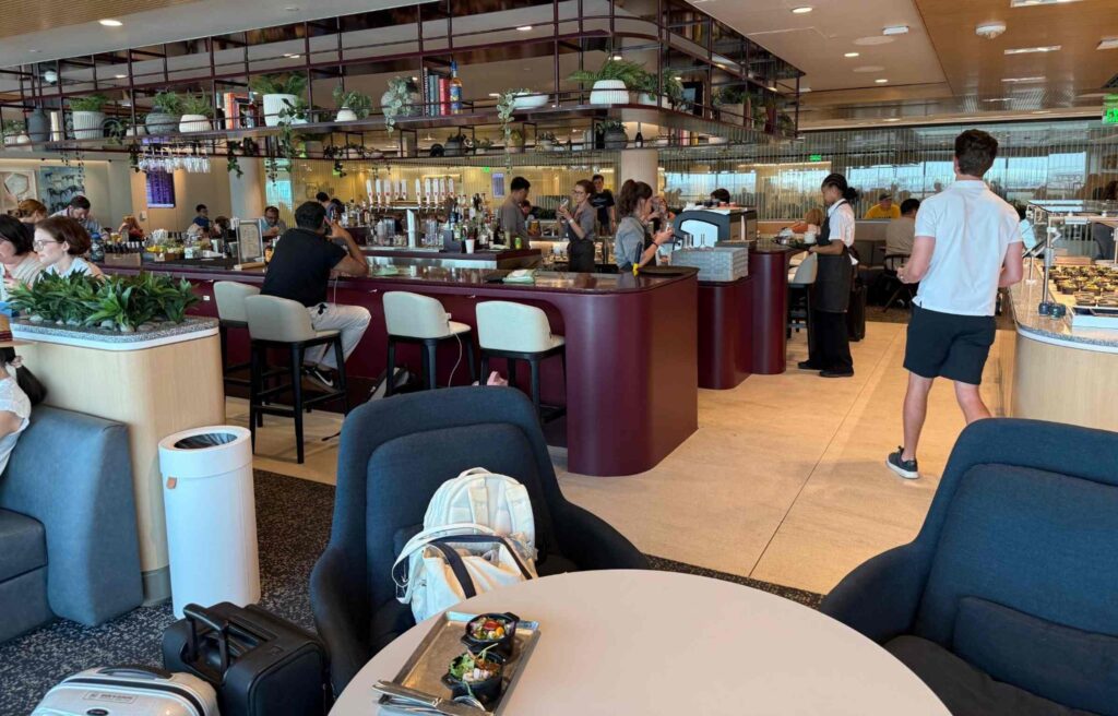 Guests relaxing in the Capital One Lounge bar and dining area, with bartenders serving drinks and travelers seated around tables and counters.