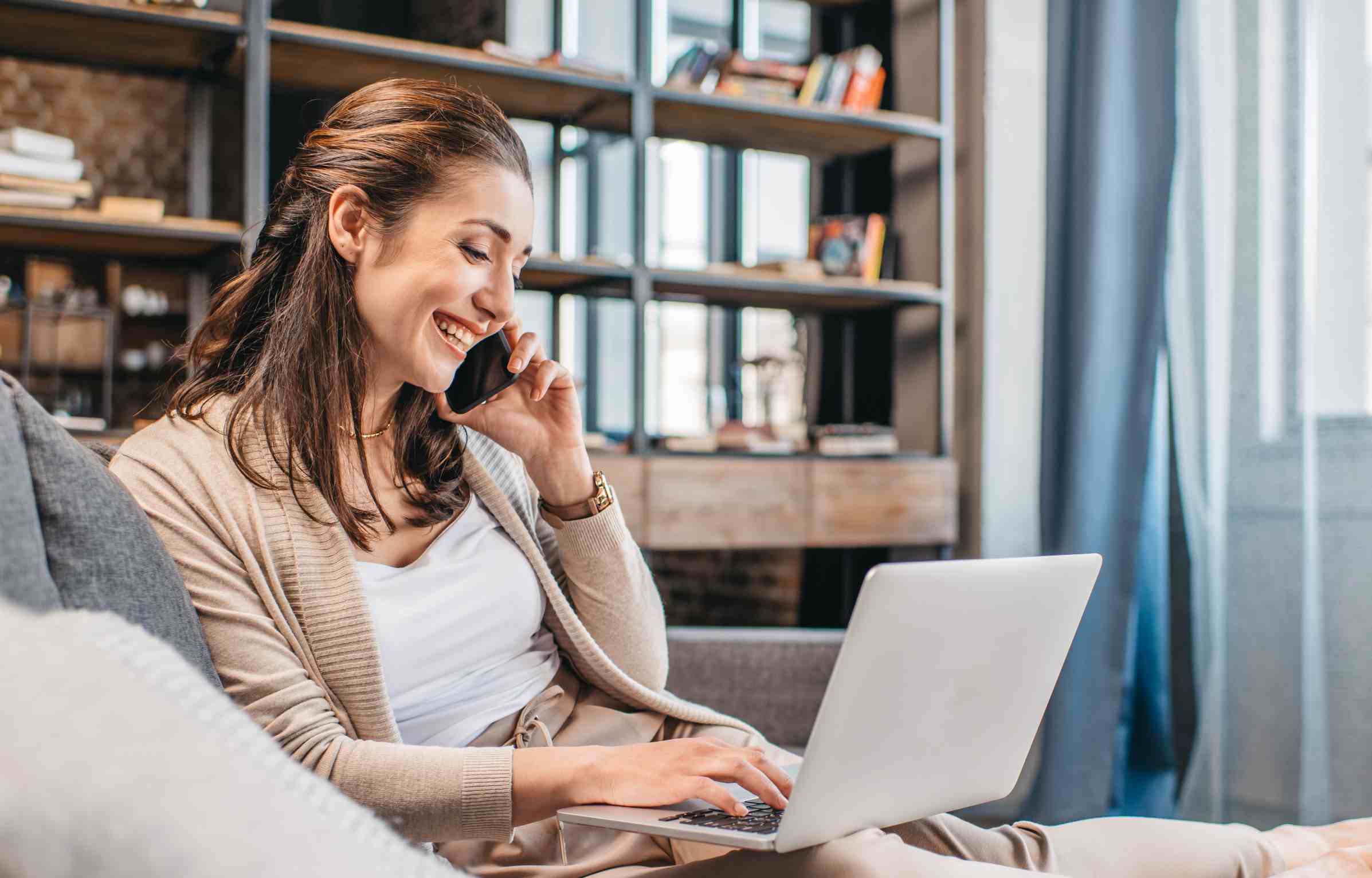 A smiling woman works remotely from home while talking on the phone and using a laptop in a cozy living room with bookshelves in the background.