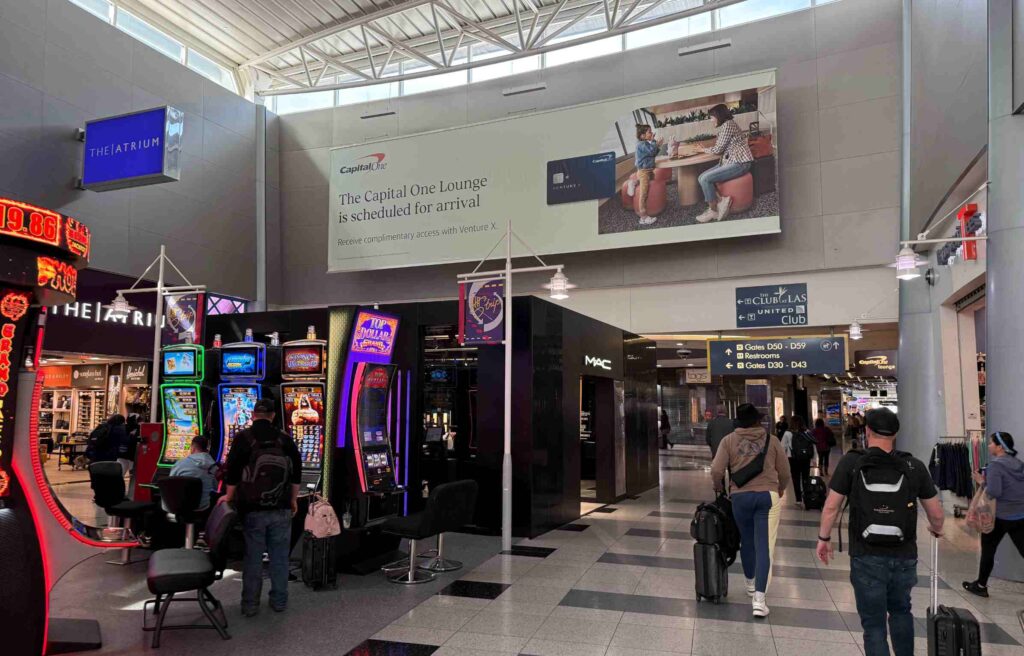 Airport corridor showing Capital One Lounge advertisement beside shops and slot machines.