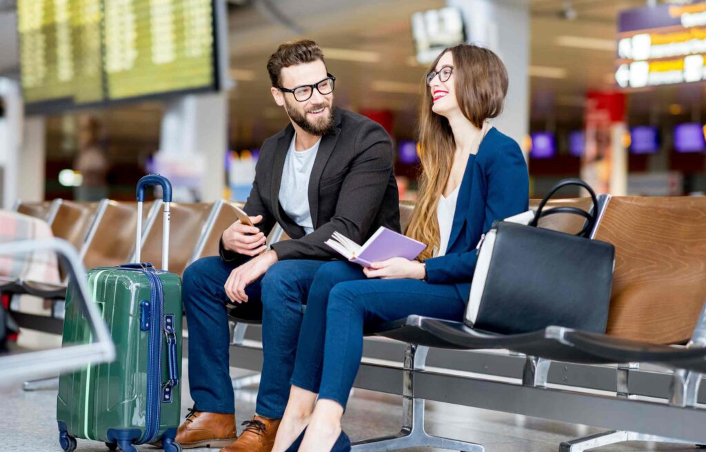 A smiling couple sitting at the airport lounge with luggage and reading before the flight, showing how to use american airlines aadvantage miles for travel.
