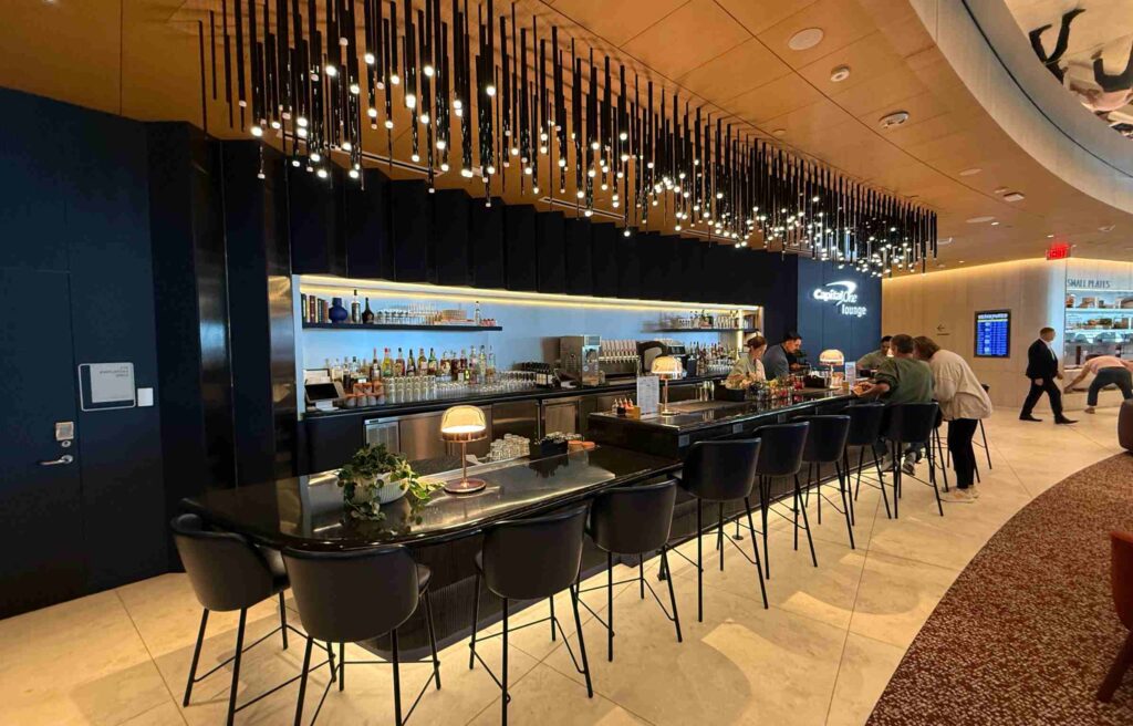 Capital One Lounge bar with hanging lights, guests seated, and staff preparing drinks.