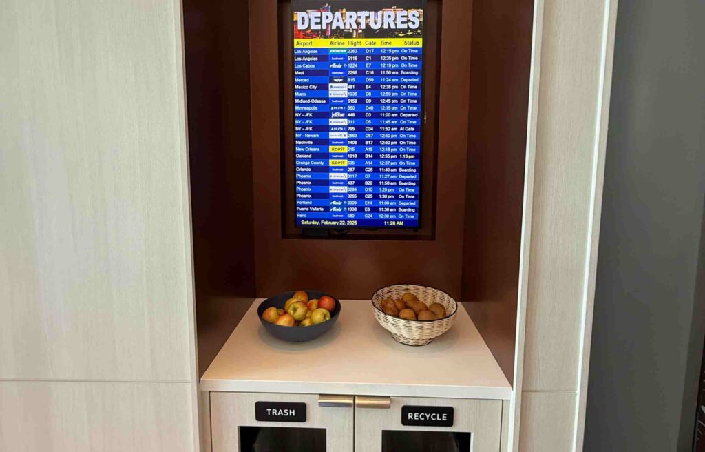 Digital departure board above baskets of apples and pears beside labeled trash and recycle bins.