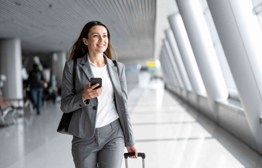 Smiling business traveler at the airport holding a phone and luggage, representing the best airline rewards program for international travel.