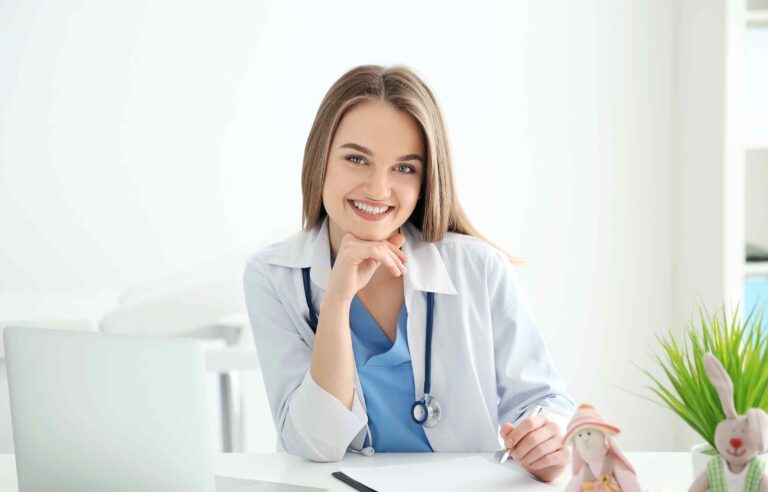 Happy female physician smiling at her desk with a stethoscope around her neck, sitting in a bright medical office.