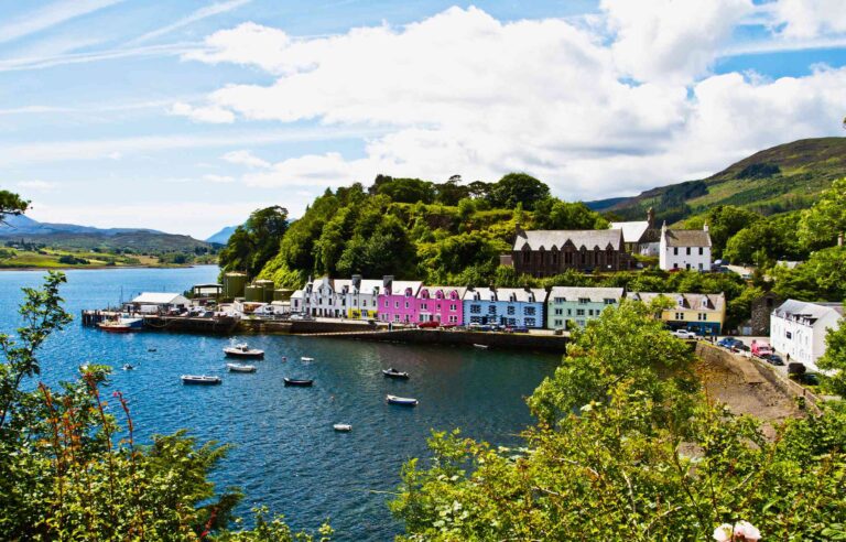 Colorful houses along Portree harbor on the Isle of Skye, Scotland, with calm water and green hills under clear skies.