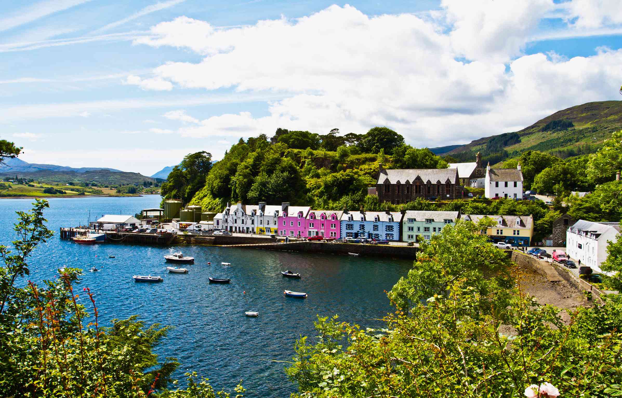 Feature image Colorful houses along Portree harbor on the Isle of Skye, Scotland, with calm water and green hills under clear skies.