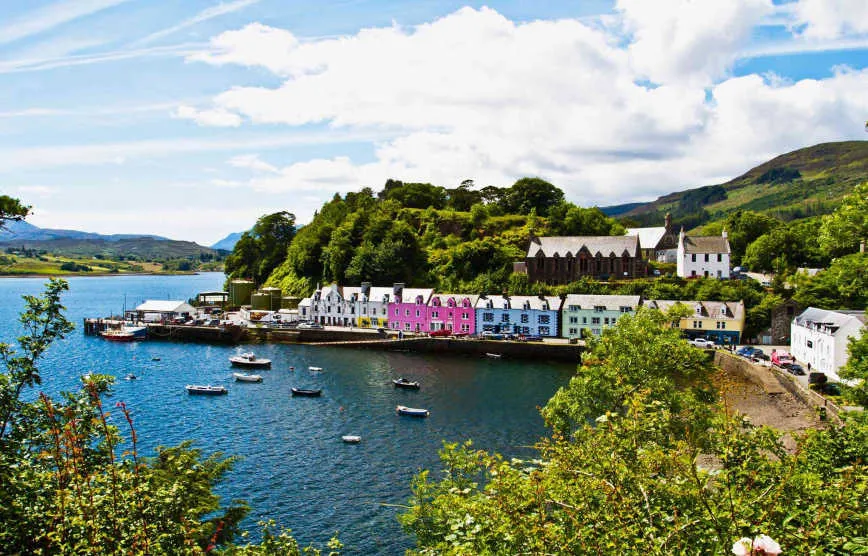 Colorful houses along Portree harbor on the Isle of Skye, Scotland, with calm water and green hills under clear skies.