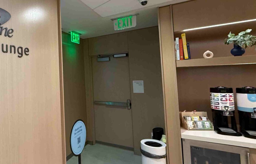 Exit area of the Capital One Lounge with beverage dispensers and shelves decorated with books and plants.