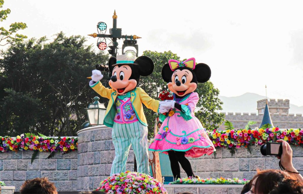 Mickey and Minnie in colorful outfits waving to a crowd during a Disney parade with bright flowers.