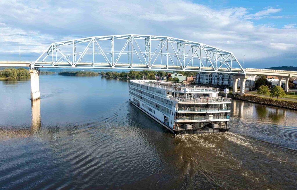 A riverboat cruising under a steel bridge on the Mississippi River near downtown Natchez, Mississippi, with waterfront homes and clear skies in the background.