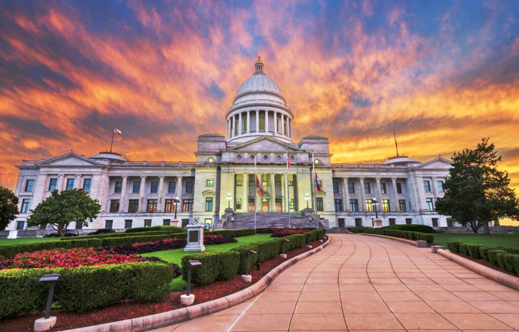 The Arkansas State Capitol building in Little Rock at sunset, with a vibrant sky and landscaped gardens, representing the growing region of Northwest Arkansas.