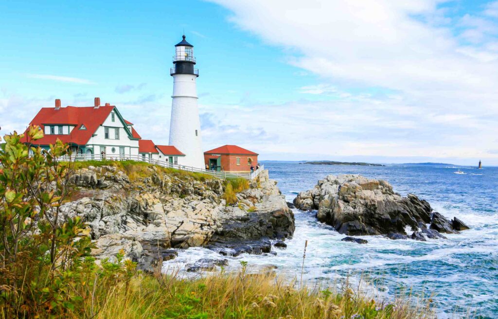 The Portland Head Light lighthouse in Portland, Maine, overlooking rocky cliffs and the Atlantic Ocean under a bright blue sky.