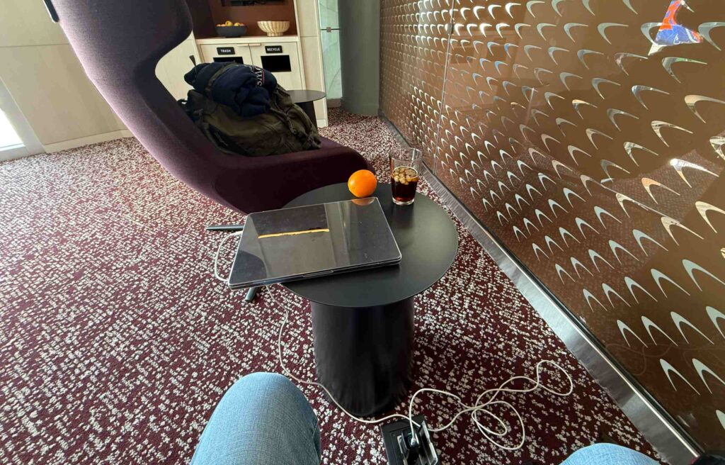 Lounge seating area with a table, drink, orange, and laptop near a patterned metallic wall.