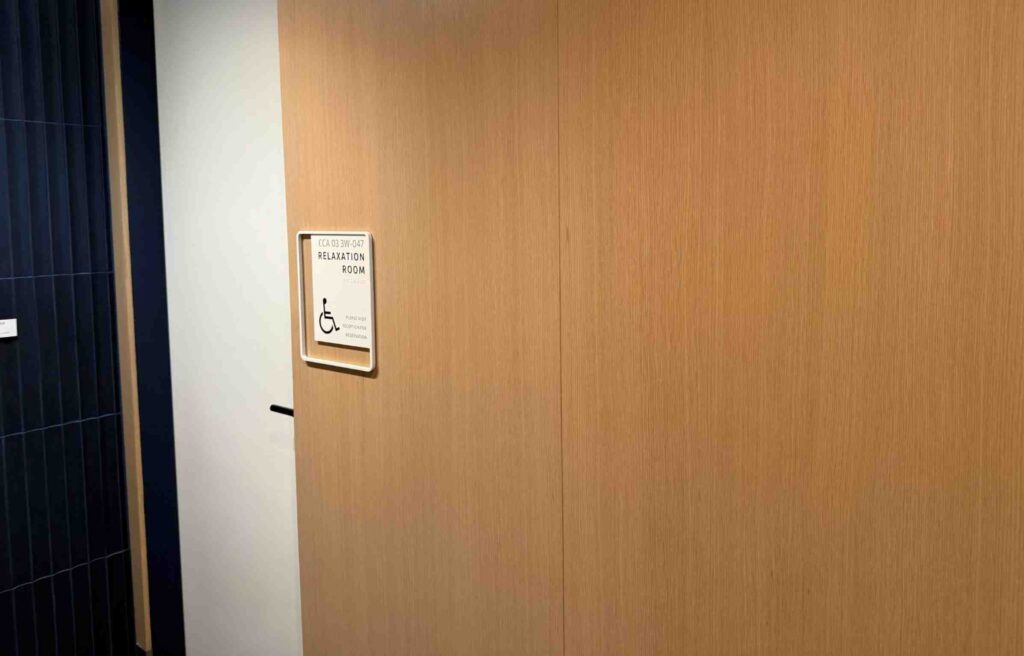 Doorway to the Capital One Lounge relaxation room with an accessibility sign and light wood walls.