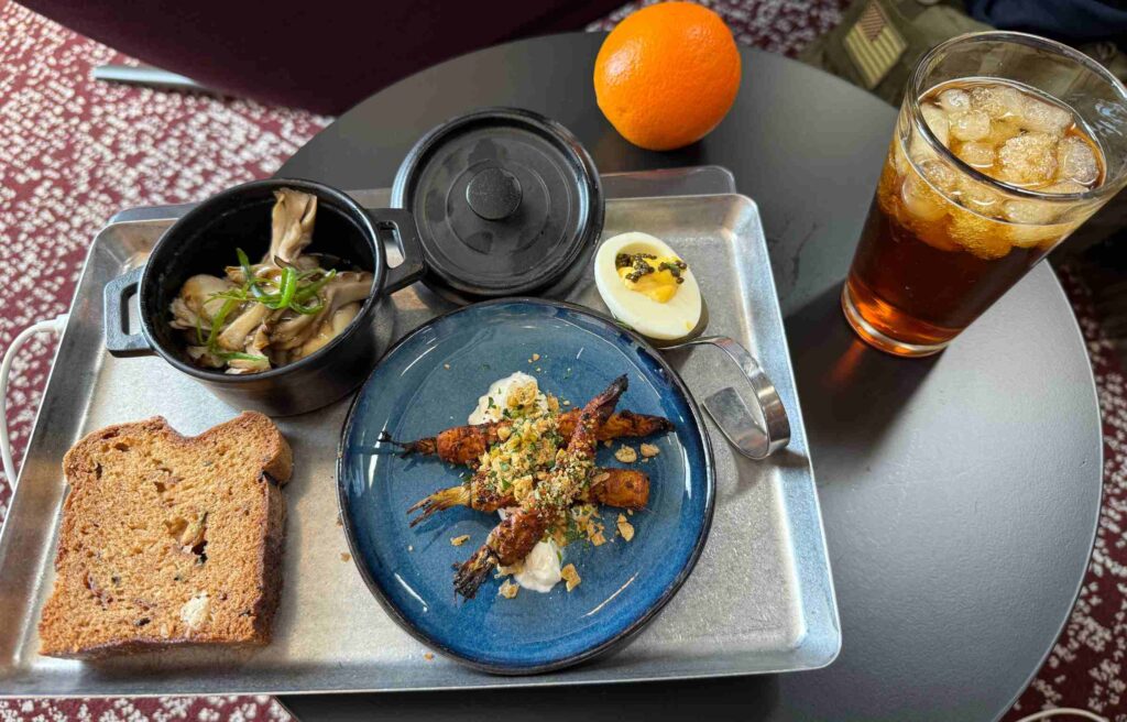 Meal tray featuring roasted carrots, caviar deviled egg, zucchini bread, and iced drink.