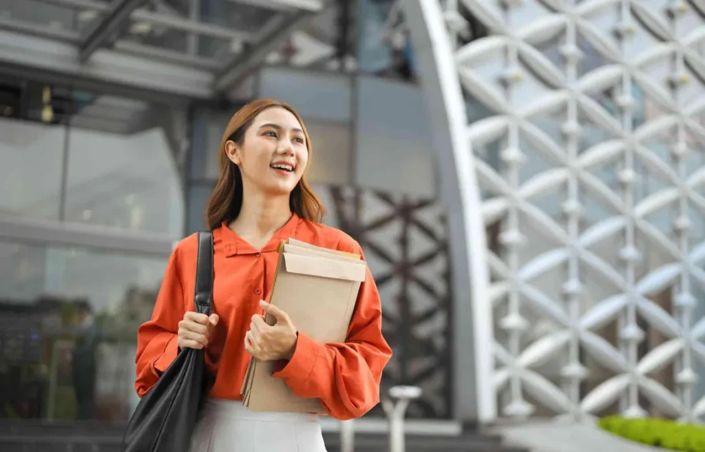 A young woman in an orange shirt smiling outside a modern office building, holding folders and a bag, representing talent relocation and global career opportunities.