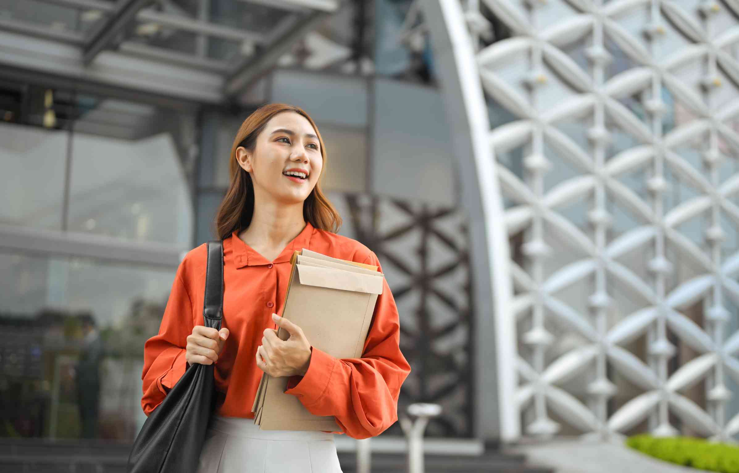 Feature image A young woman in an orange shirt smiling outside a modern office building, holding folders and a bag, representing talent relocation and global career opportunities.