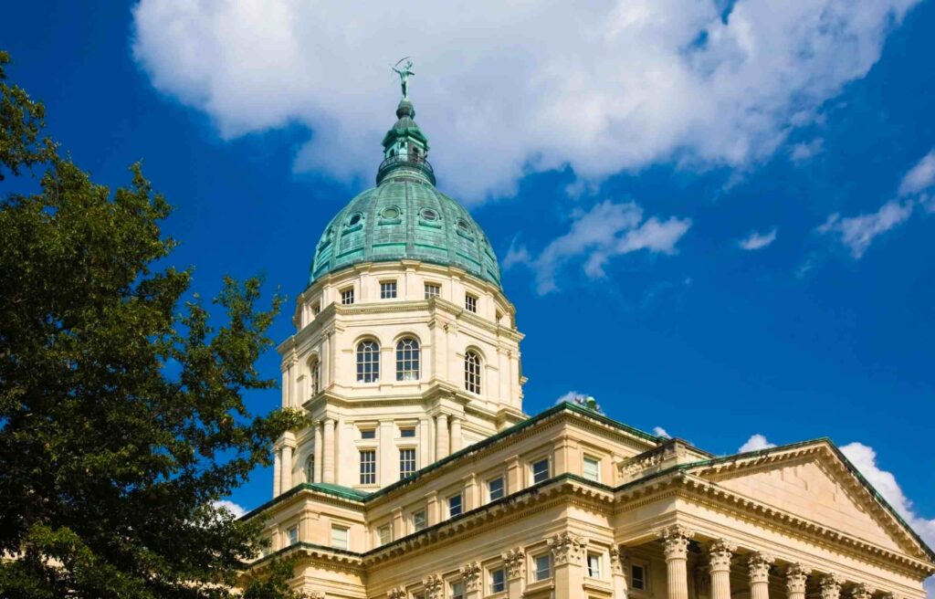 The Kansas State Capitol building in Topeka, featuring its green dome under a bright blue sky with scattered clouds.