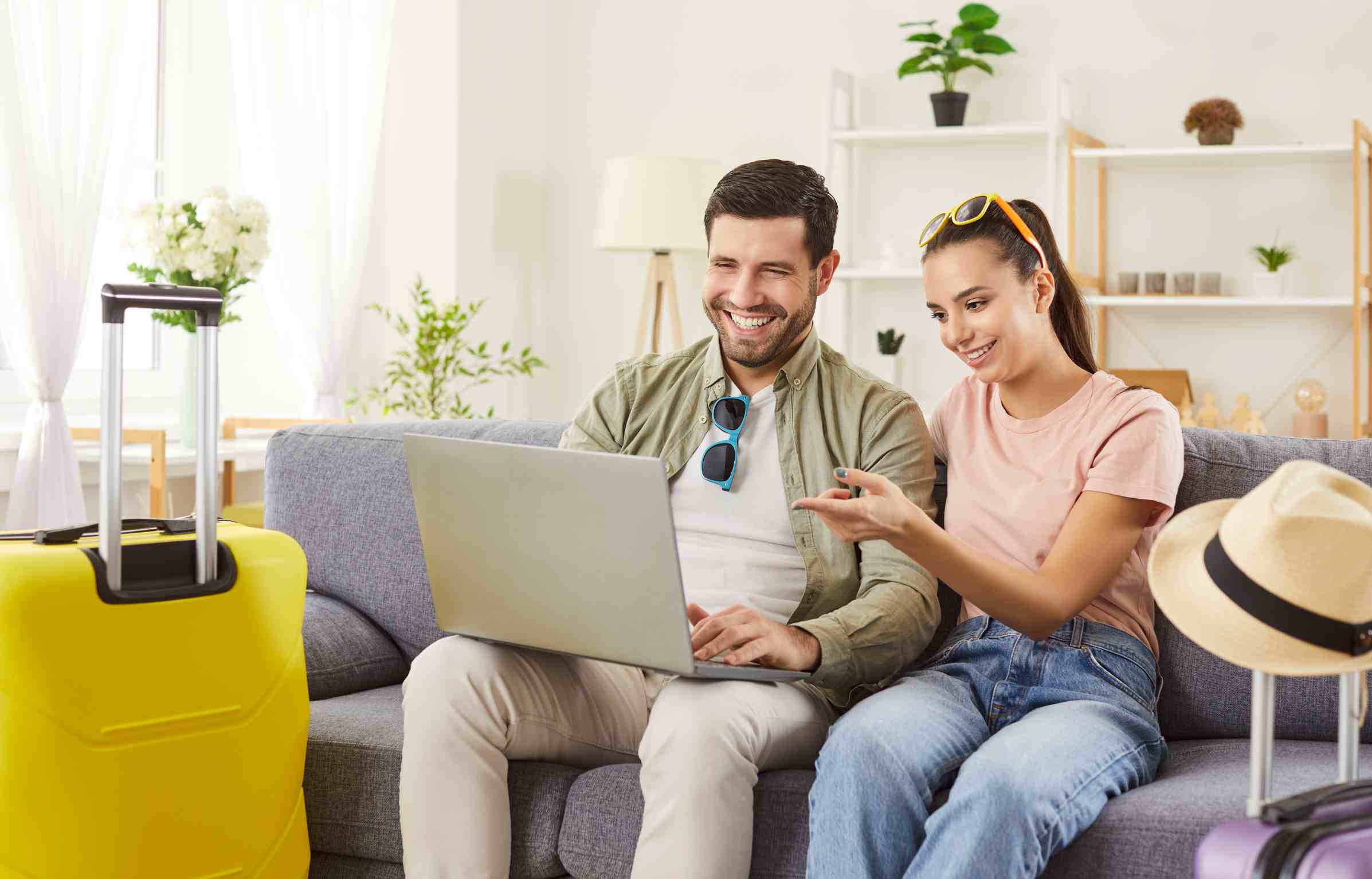 A smiling couple sitting on a couch with suitcases nearby, using a laptop to book flights and hotels online.