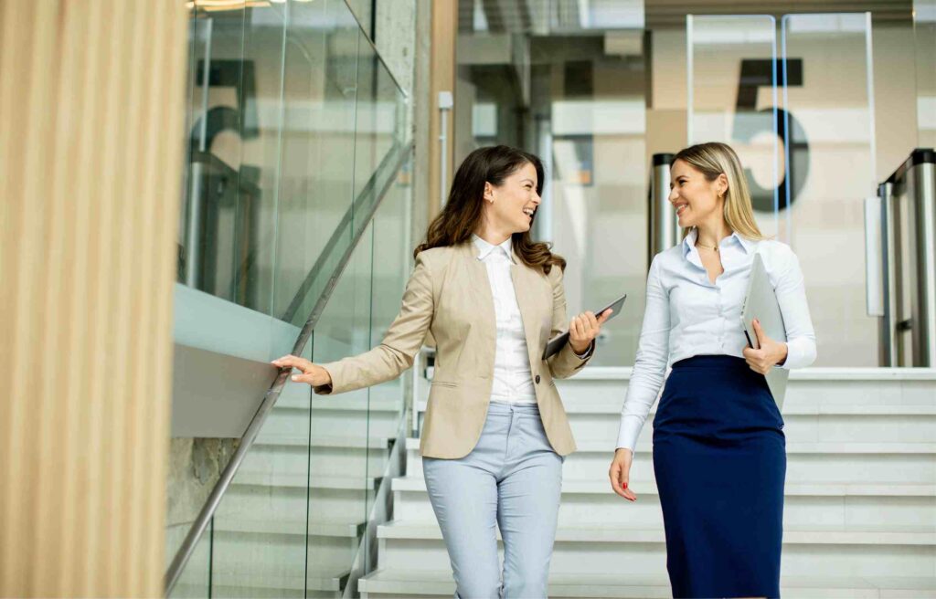 Two women walking and talking inside a modern office building, holding laptops and tablets, representing teamwork and talent relocation.