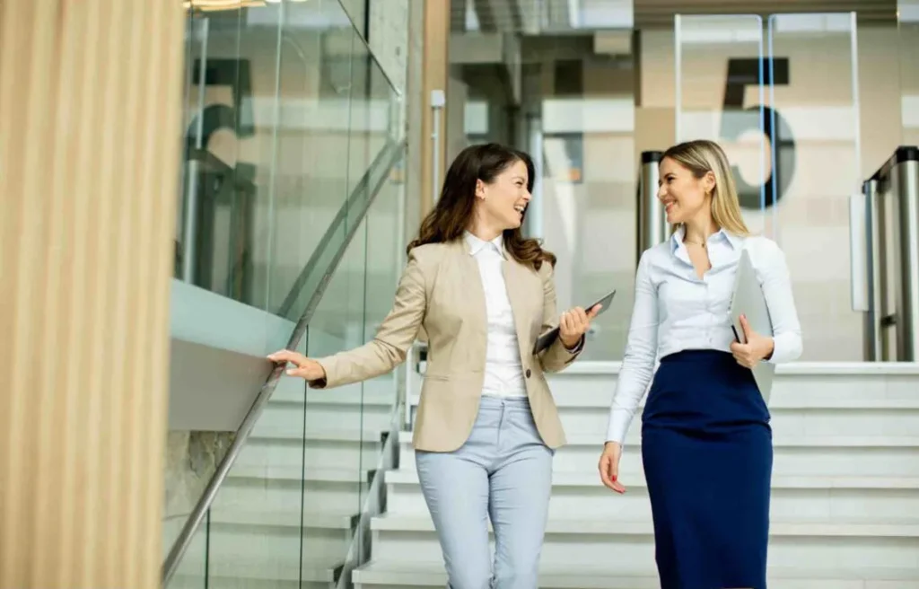 Two women walking and talking inside a modern office building, holding laptops and tablets, representing teamwork and talent relocation.