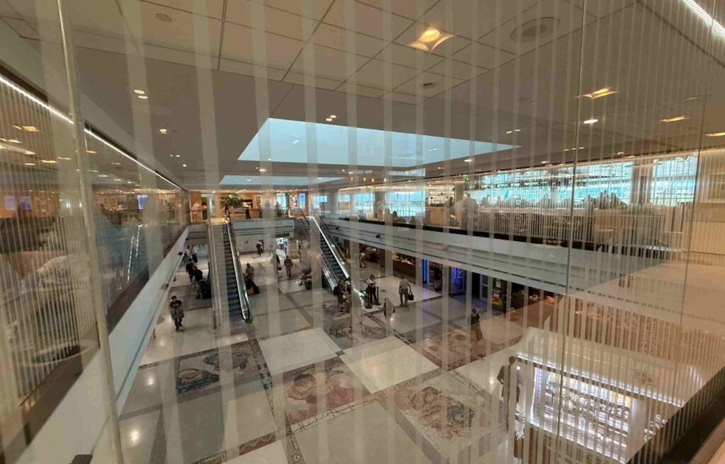 Wide view from the Capital One Lounge overlooking the concourse at Denver Airport, showing escalators and travelers below.