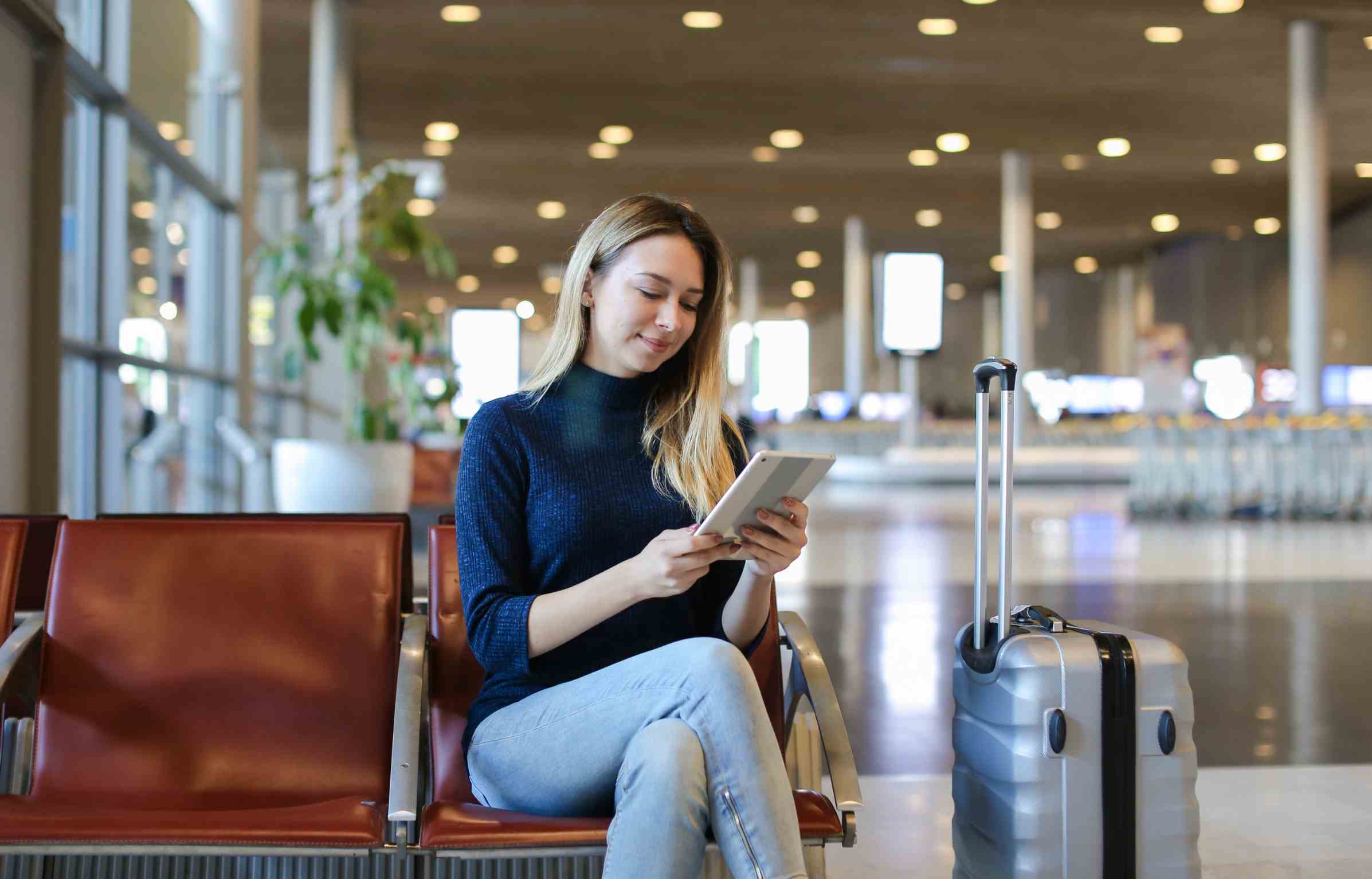 A woman sitting at an airport terminal with her suitcase beside her, checking flight compensation details on a tablet while waiting for departure.