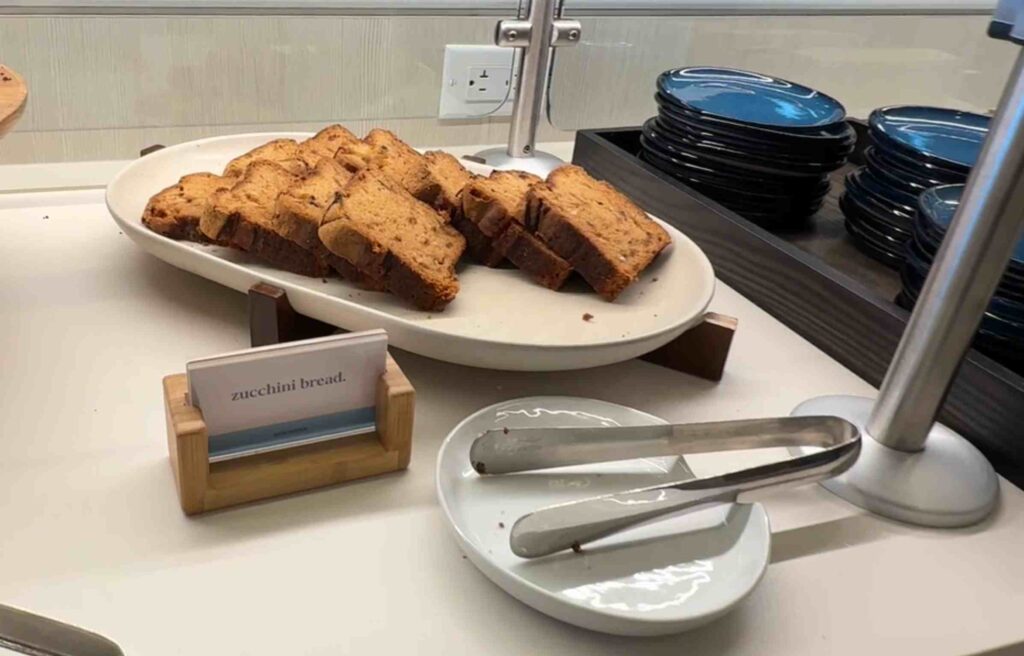Sliced zucchini bread arranged on a white platter beside small serving plates and tongs.