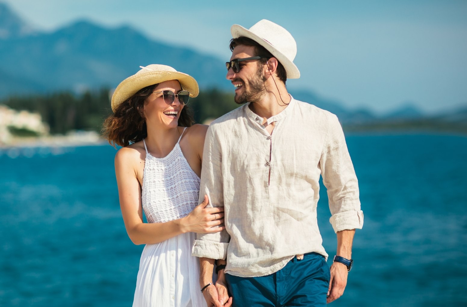 Joyful couple walking hand in hand along the waterfront, dressed in summer outfits and smiling warmly.