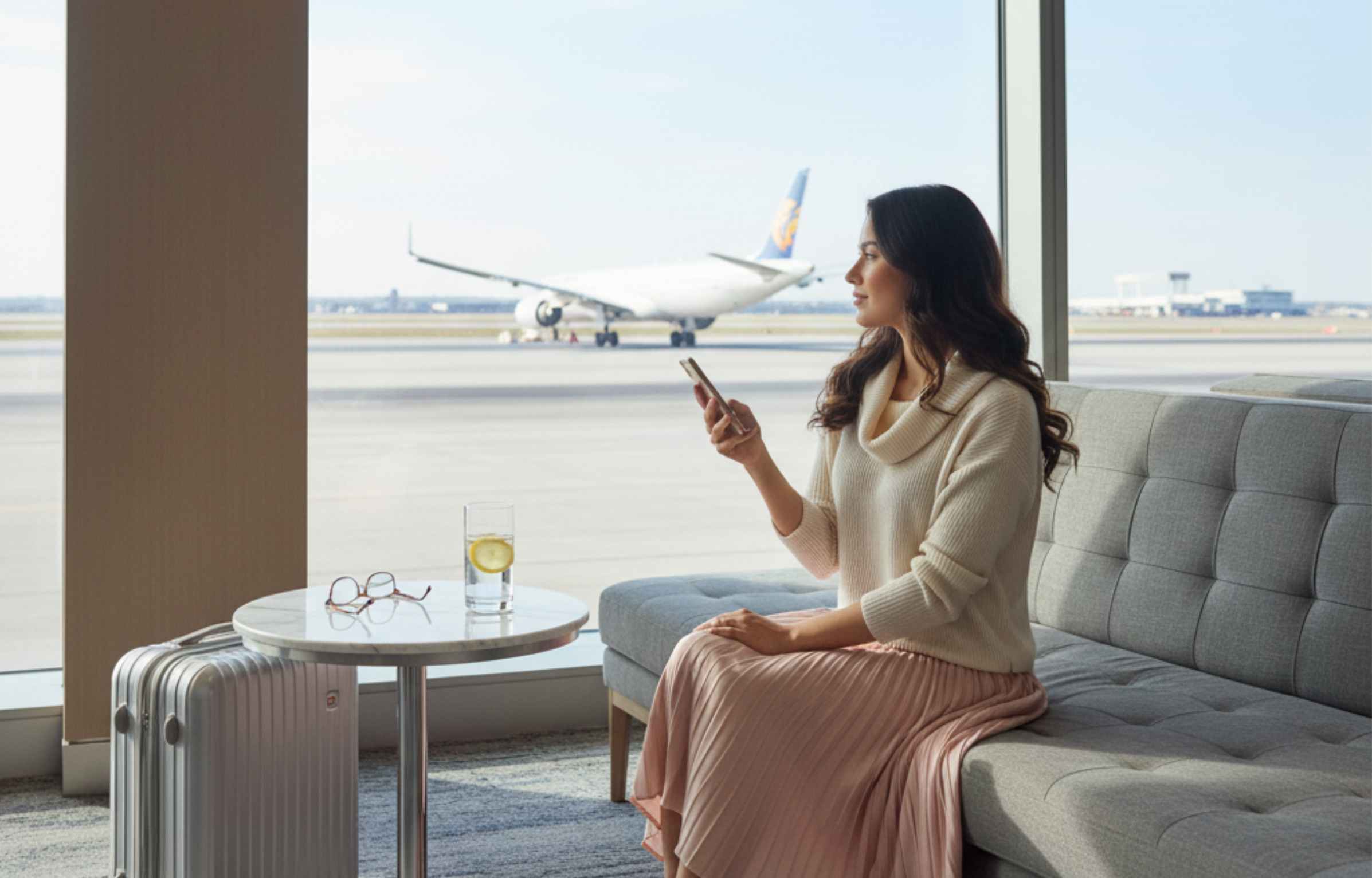 Traveler sitting in airport lounge near window, holding phone and watching plane on runway.