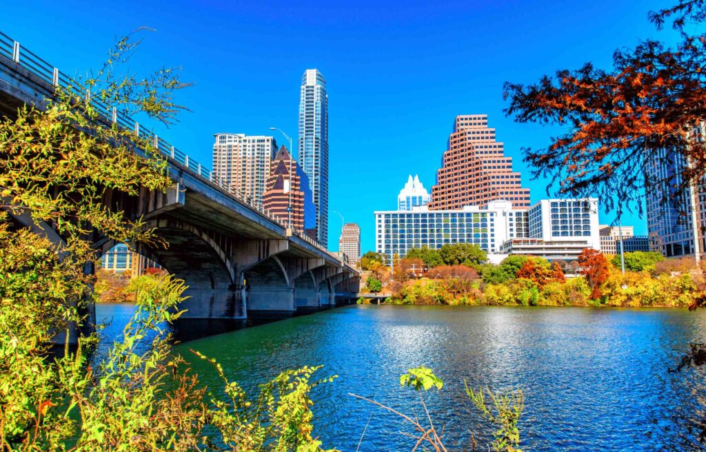 Downtown Austin, Texas skyline with modern buildings viewed across Lady Bird Lake, framed by a bridge and colorful autumn trees under a clear blue sky.
