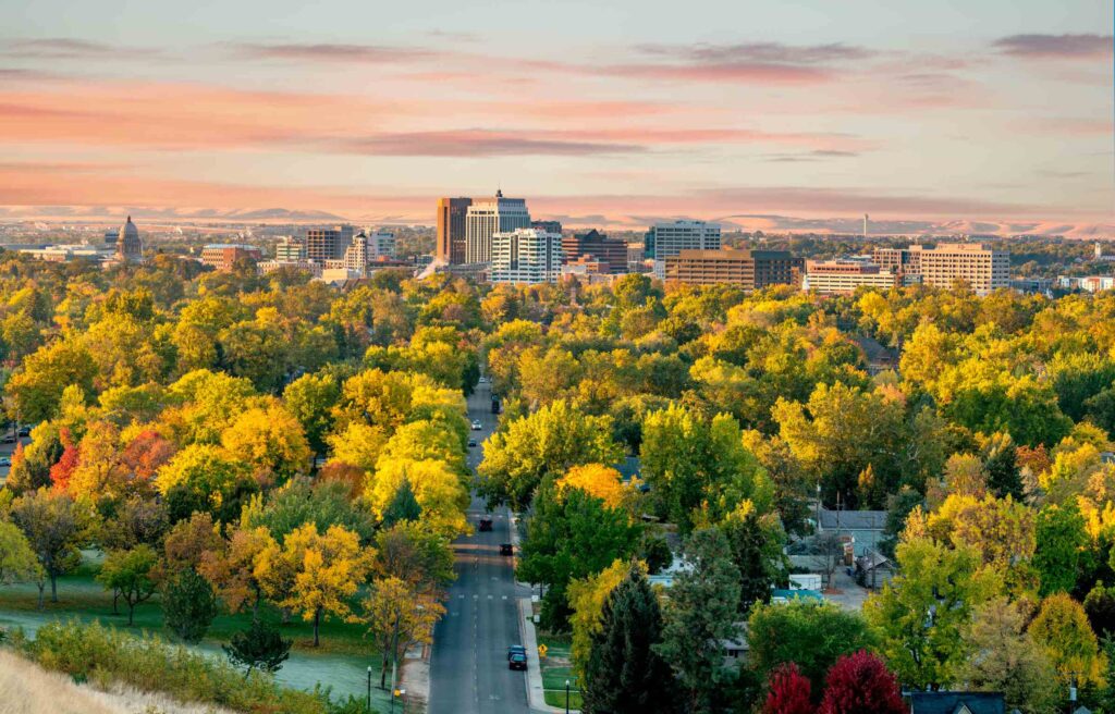 Downtown Boise, Idaho skyline surrounded by colorful autumn trees, with the Capitol building visible under a soft orange and pink sunset.