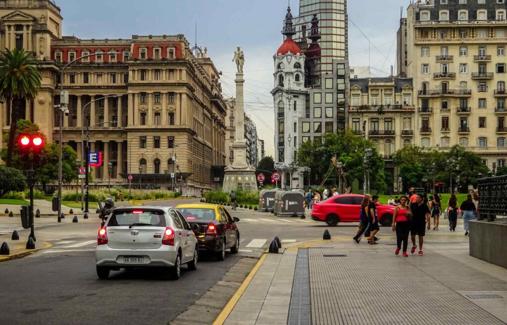 A city scene in Buenos Aires, Argentina, showing historic buildings, a central monument, cars, and pedestrians at a busy intersection.