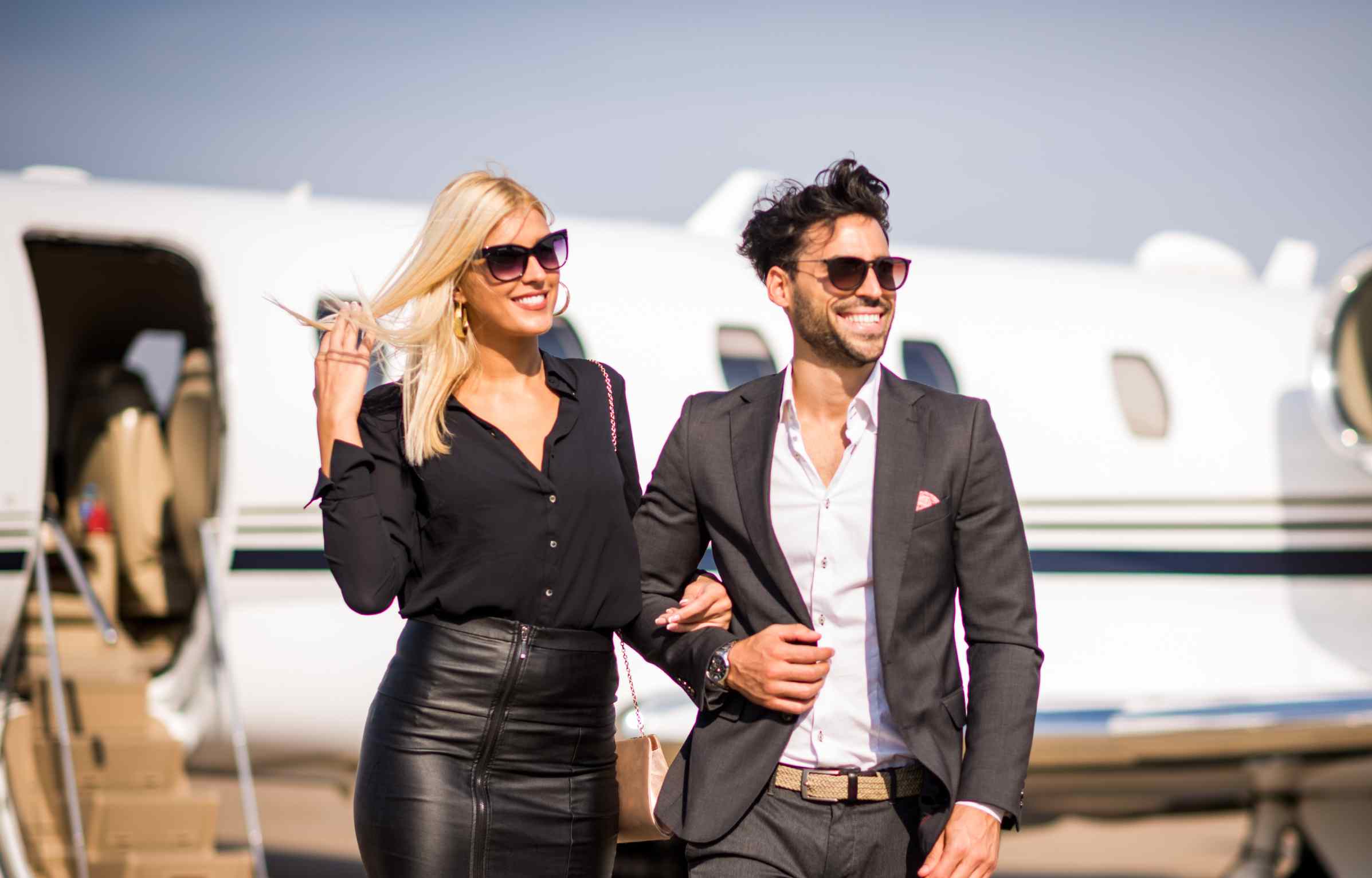 A well dressed couple walks away from a private jet, both wearing sunglasses and smiling, with the aircraft staircase visible in the background.