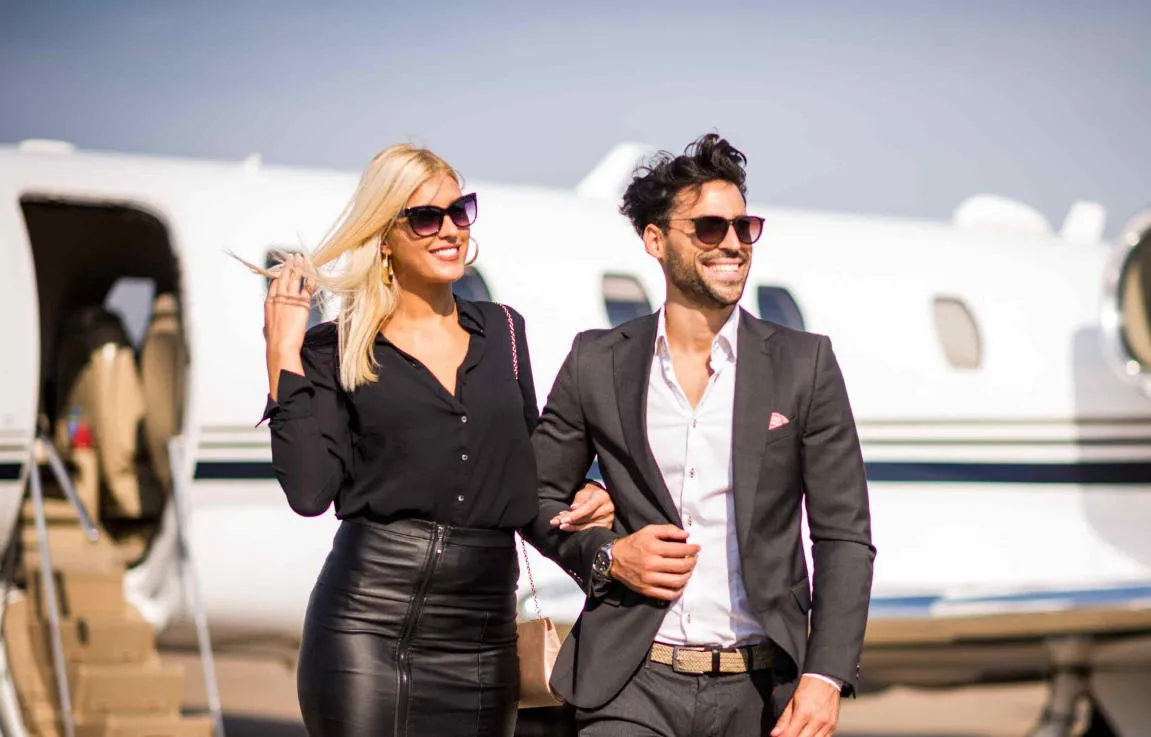 A well dressed couple walks away from a private jet, both wearing sunglasses and smiling, with the aircraft staircase visible in the background.
