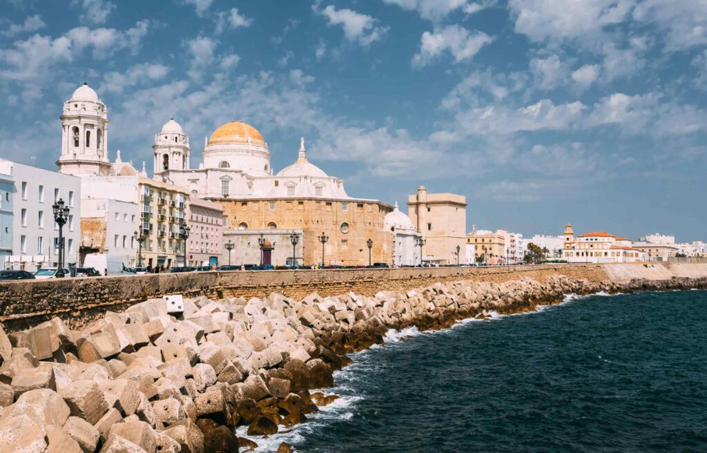 The Cádiz Cathedral and seafront in Cádiz, Spain, with waves hitting the rocky shoreline under a bright sky.
