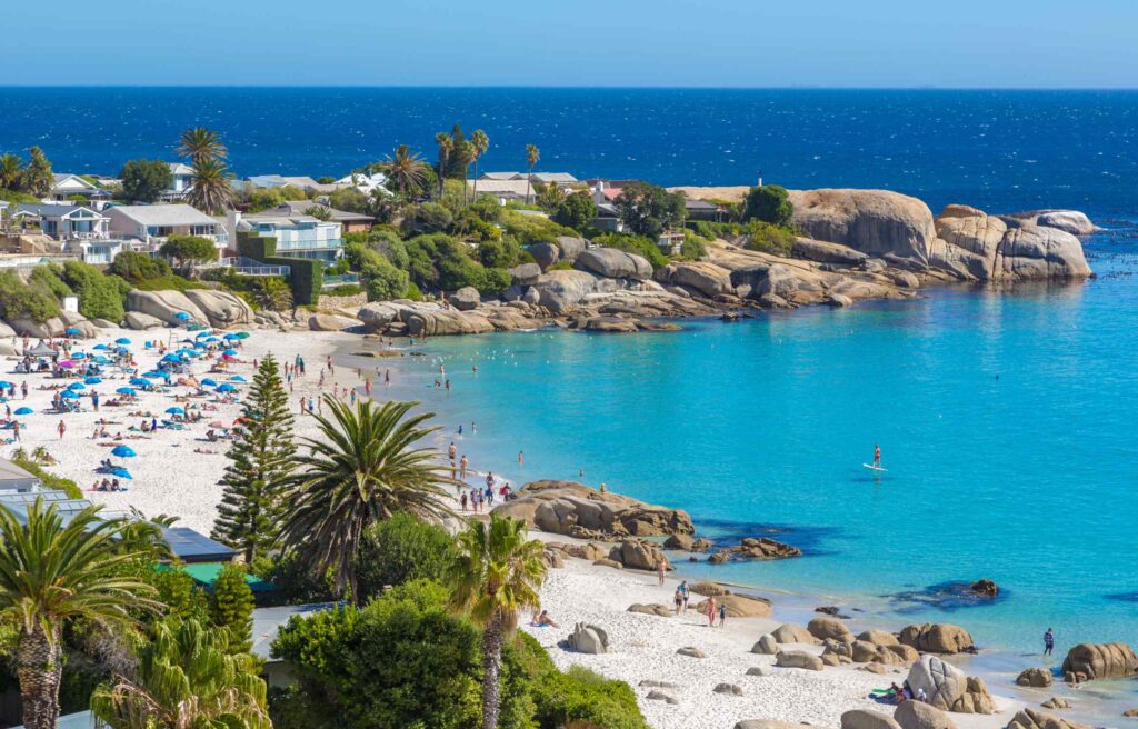 A scenic beach in Cape Town, South Africa, with turquoise water, white sand, rocky cliffs, and people enjoying the shoreline.
