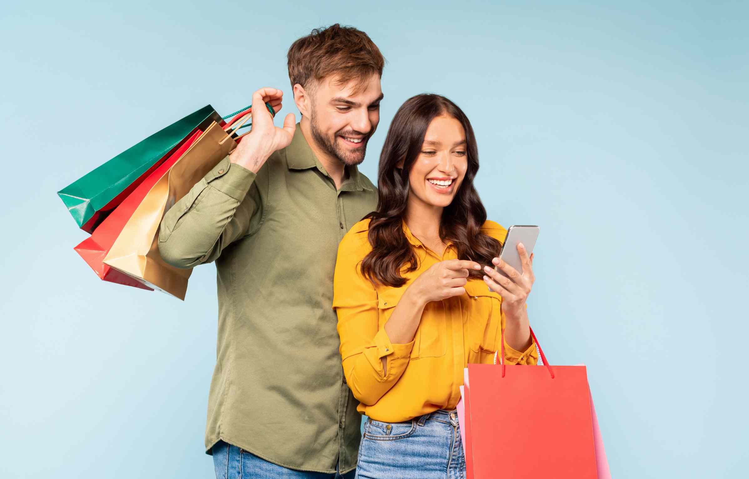 Couple smiling while checking a smartphone and carrying colorful shopping bags against a light blue background.