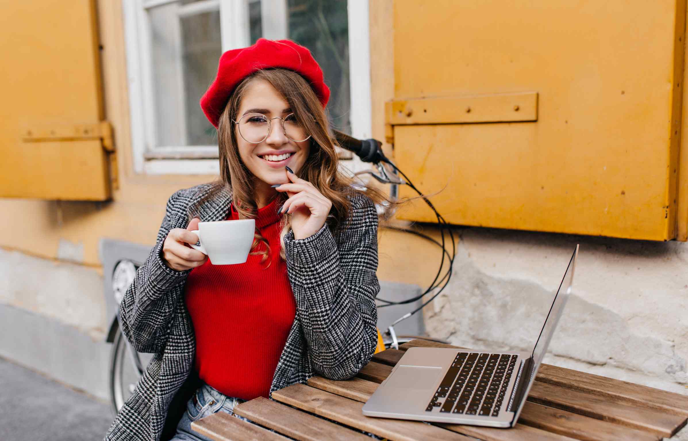 A smiling woman in a red beret sits at an outdoor café with a laptop and a cup of coffee.