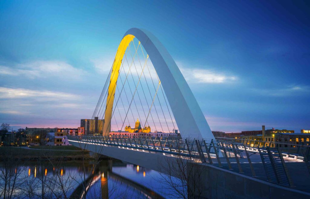 The illuminated Iowa Women of Achievement Bridge in Des Moines at dusk, reflecting over the Des Moines River with the city skyline in the background.