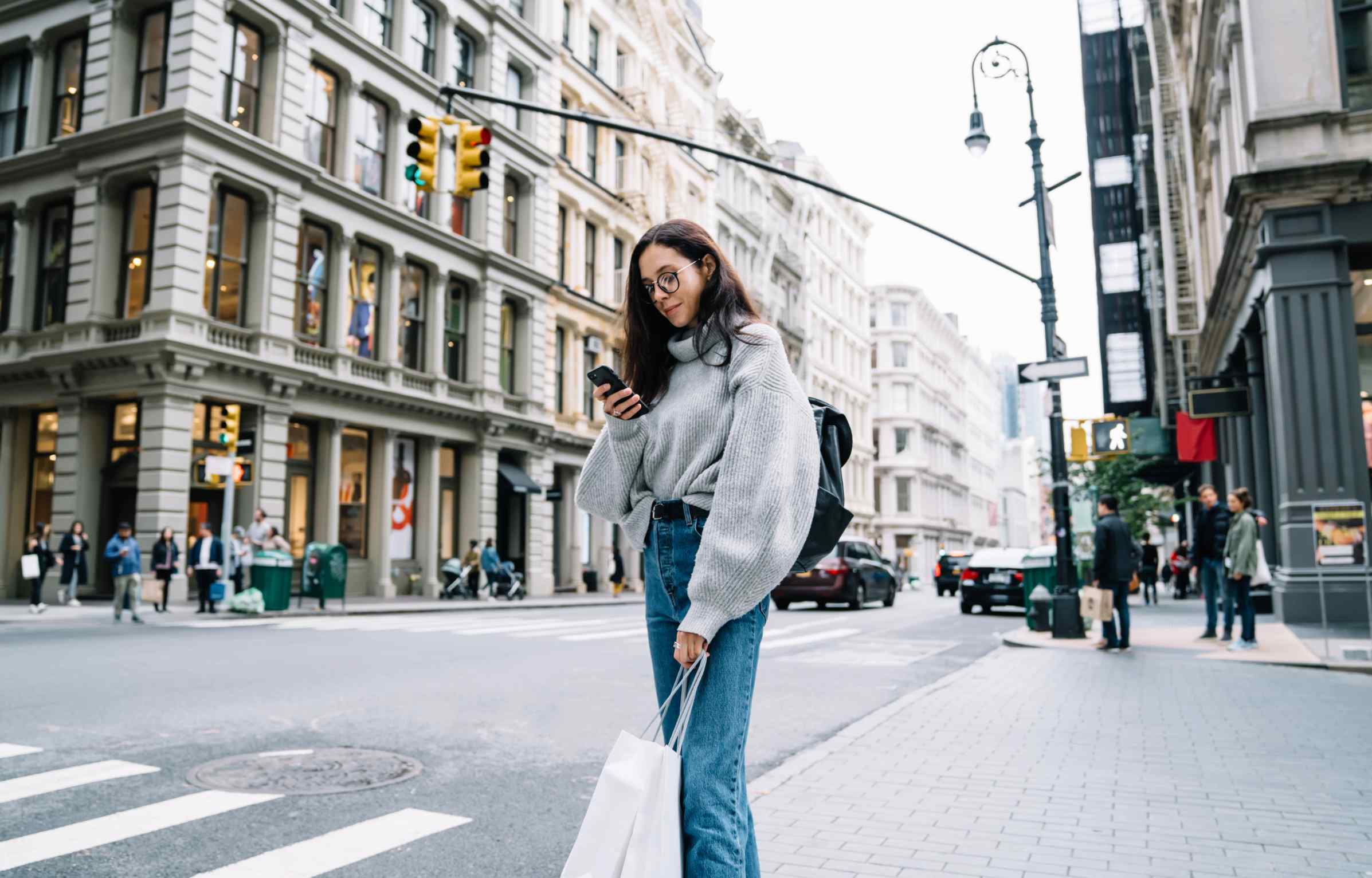 A young woman stands at a city crosswalk checking her phone, holding shopping bags, surrounded by historic buildings and light traffic.